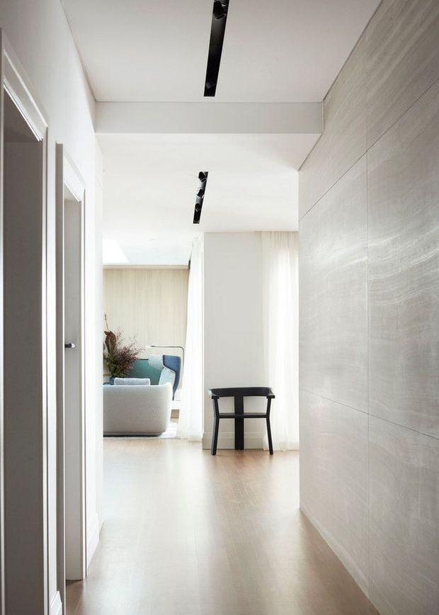 Bright minimalist hallway with pale timber flooring, white walls and ceiling, a textured light-toned feature wall, slim black recessed linear lighting, and doorways along the left side. The hallway opens into a softly furnished living room with a pale sofa and a small black console or bench.