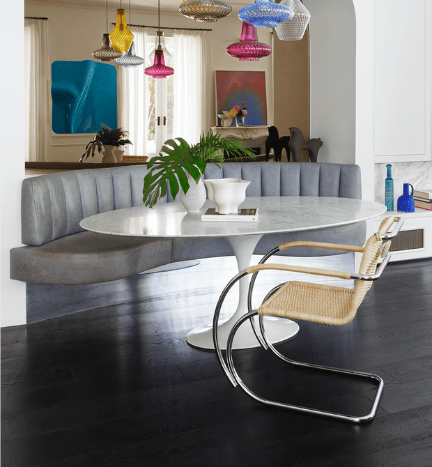 A refined dining area with a curved grey upholstered banquette, white oval pedestal dining table, and a cantilevered woven-and-chrome dining chair. The room features dark timber flooring, tall sheer-draped windows, colourful sculptural pendant lights, contemporary artwork, indoor greenery, and white built-in cabinetry with a marble-look surface. The overall composition is polished, artistic, and contemporary with mid-century-inspired furniture accents.