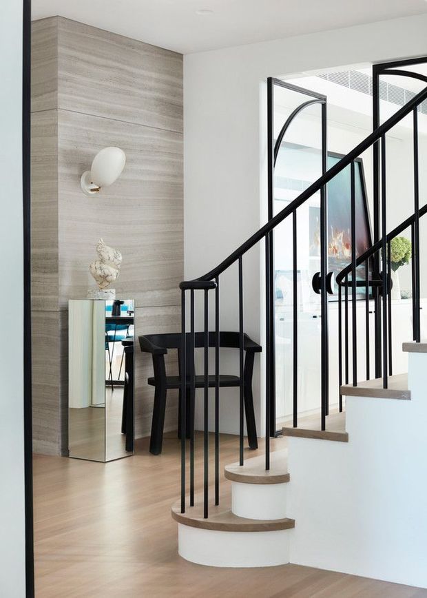 Bright contemporary stairway and entry area featuring a curved white staircase with light timber treads, slim black metal balustrades, pale timber flooring, and a vertically panelled stone or timber-look feature wall. A black armchair, mirrored console, sculptural decor, and glazed interior doors are visible.