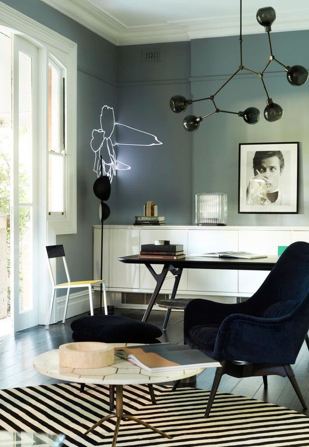 A refined home office with dark blue-grey walls, decorative wall panelling and classic crown moulding. The room includes a contemporary black desk, sculptural black pendant light, white built-in cabinetry, a framed artwork, a sculptural floor lamp, lounge chair with ottoman, and a round coffee table over a bold black-and-white striped rug. Tall white-framed windows provide abundant natural light.