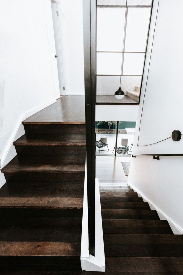 A contemporary interior stairway with dark timber stair treads, crisp white walls, and a slim black-framed glazed balustrade or void divider. The stairs connect a bright upper landing with a lower living area visible through the opening, featuring a large skylight or glazed roof and a black pendant light. The composition emphasizes vertical circulation, strong contrast, and clean architectural detailing.