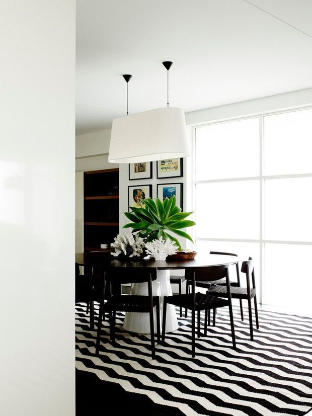 Bright dining room with a dark timber dining table and chairs, a large white pendant light, a bold black-and-white wavy patterned rug, framed artwork, dark built-in shelving, and a large indoor plant arrangement. Expansive windows provide abundant natural light.