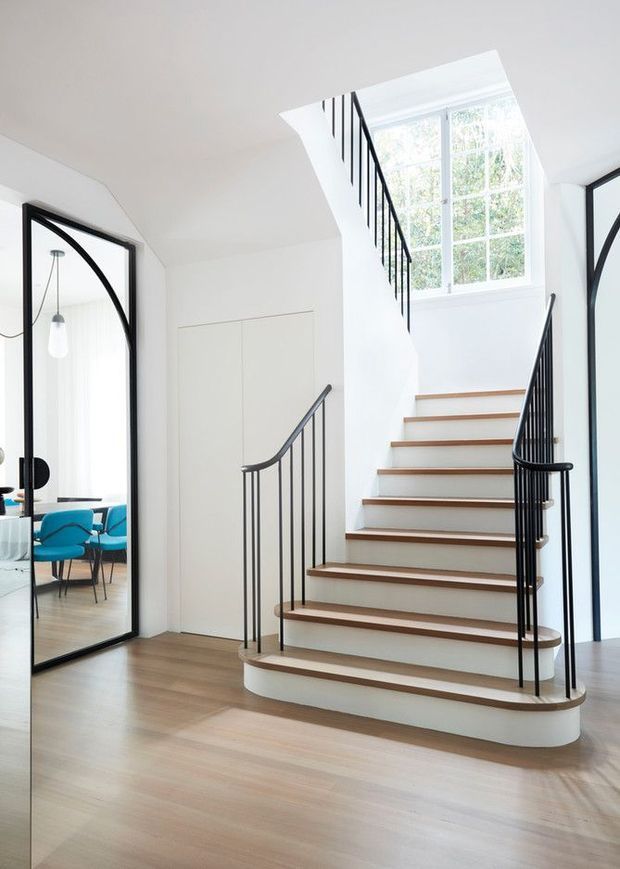 Bright contemporary stairway with curved, white-painted stair risers and light timber treads. Slim black metal balustrades frame the staircase, which rises beside a tall multi-pane window and an internal glazed doorway. Light timber flooring, white walls, and glimpses of a colourful sitting area create a clean, refined interior.