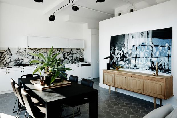 Open-plan contemporary kitchen and dining area with white cabinetry, a dramatic black-and-white stone splashback, dark dining table and chairs, and a timber sideboard beneath a large artwork or television. The space features distinctive hexagonal floor tiles and sculptural black ceiling lighting.
