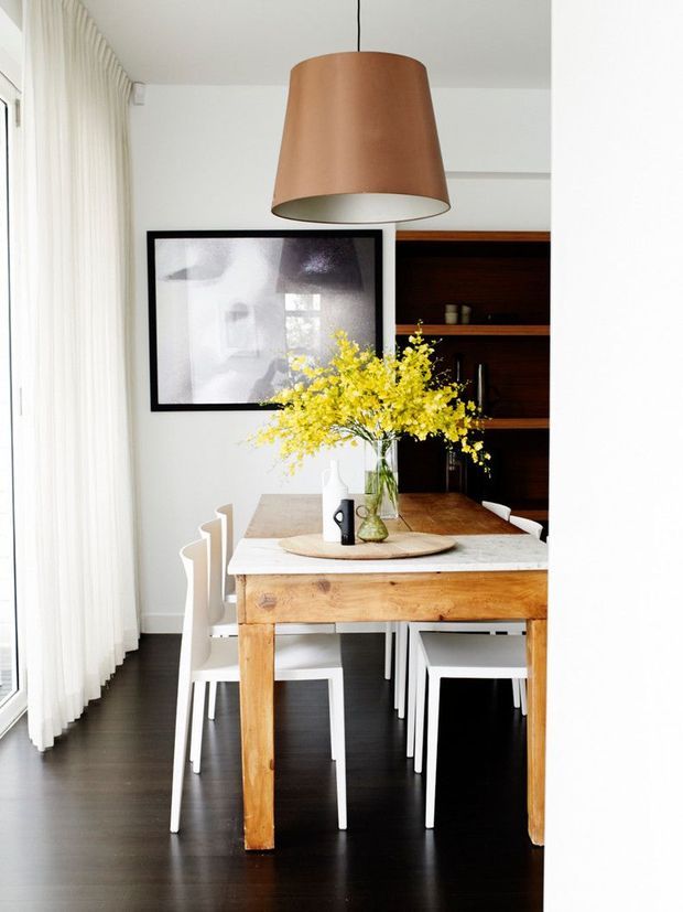 Bright, refined dining area with a rustic timber dining table and white chairs, set against dark timber flooring and a built-in dark wood shelving unit. Sheer curtains filter daylight from a large window, while a large copper pendant light, framed artwork, and a vase of yellow flowers add warmth and contrast.