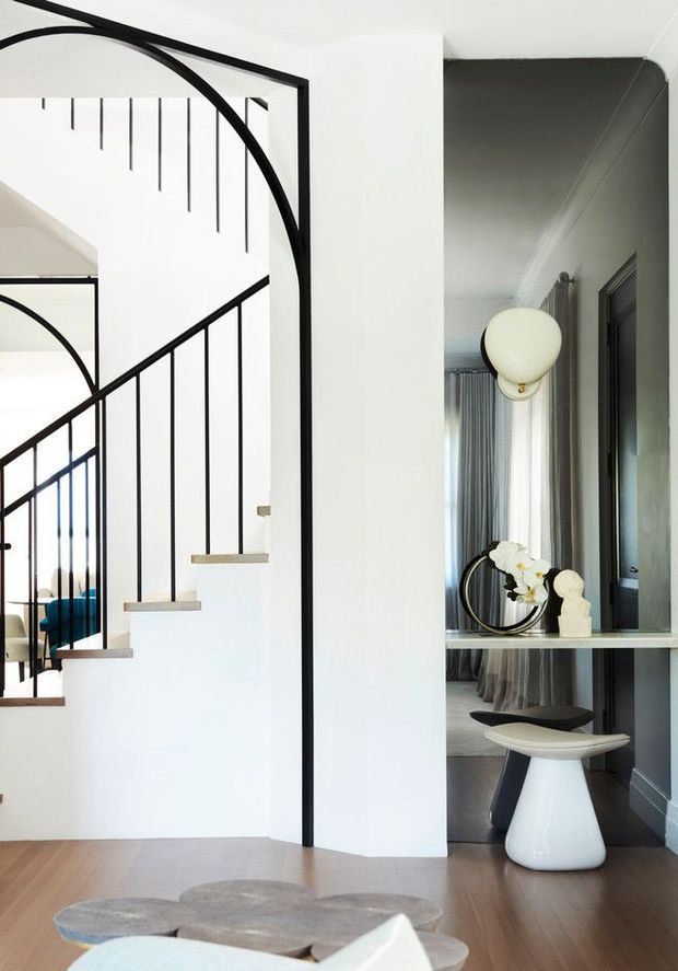 Bright contemporary entry hall featuring a sculptural black metal staircase with slender vertical balusters, pale timber treads, and a dramatic curved black-framed opening. A compact console with mirror, pendant light, decorative objects, and stool sits to the right, while a glimpse of a blue sofa is visible beyond the stair. The palette is predominantly white, charcoal grey, black, and light timber.