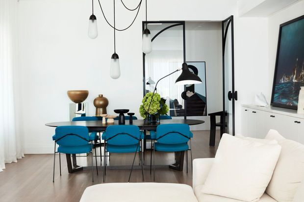 Bright open-plan dining and living area with a contemporary dining table and vivid blue upholstered chairs, white sofa, pale timber flooring, sculptural pendant lights, black-framed glazed doors, and a white built-in media cabinet. A large mirror reflects the dining and living space.