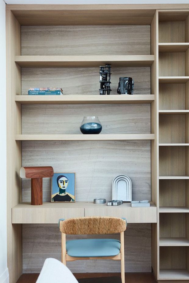 A compact built-in timber study nook with open shelving, a desk, and a chair. The cabinetry is finished in light natural wood with a wood-look back panel. Shelves display books, a camera, decorative objects, artwork, and a lamp, creating a neatly curated workspace.