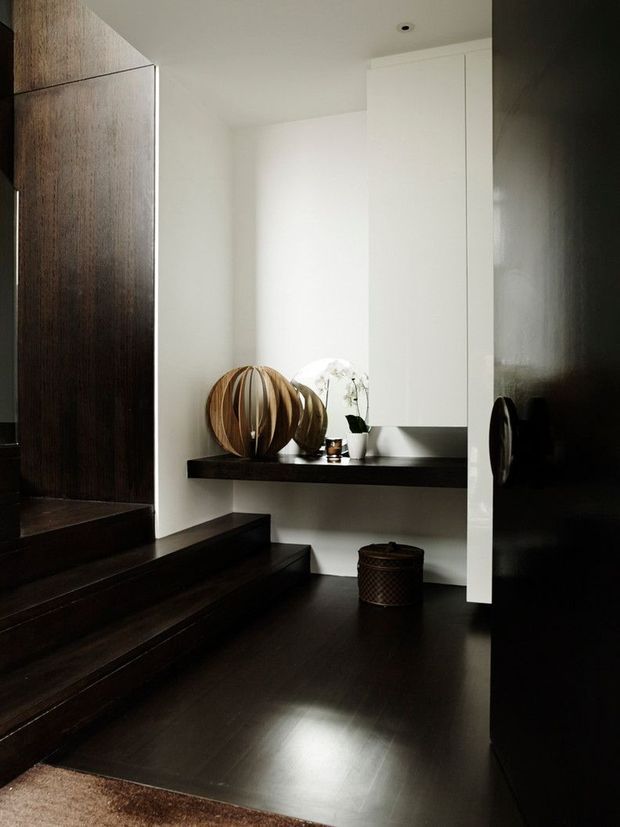 A dark, refined interior entryway with wide timber stairs, dark timber flooring and matching dark wall panelling. A floating black console shelf displays sculptural wooden objects and a small plant beneath minimalist white built-in cabinetry. The space is softly lit with a strong contrast between the white cabinetry and dark finishes.