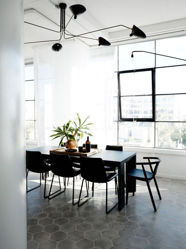 Bright contemporary dining area in a loft-like interior, featuring a black rectangular dining table with black chairs, large industrial-style windows, sheer white curtains, exposed ceiling services, sculptural black pendant lighting, and grey hexagonal floor tiles.