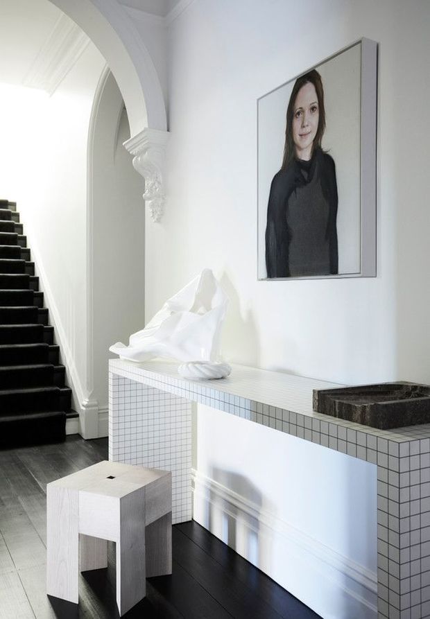 A bright, refined entry hall with a black-carpeted staircase, ornate white arched opening and column detailing, and a tiled built-in console or mantel. White sculptural decor and a framed portrait sit above the console, while a small pale timber stool stands on dark timber flooring.