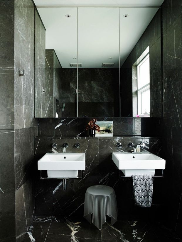 A sophisticated contemporary bathroom with twin white wall-mounted basins, dark veined marble-look wall and floor surfaces, a large mirrored wall, chrome tapware, and a high side window. The space has a dramatic black-and-white palette and compact double-vanity layout.