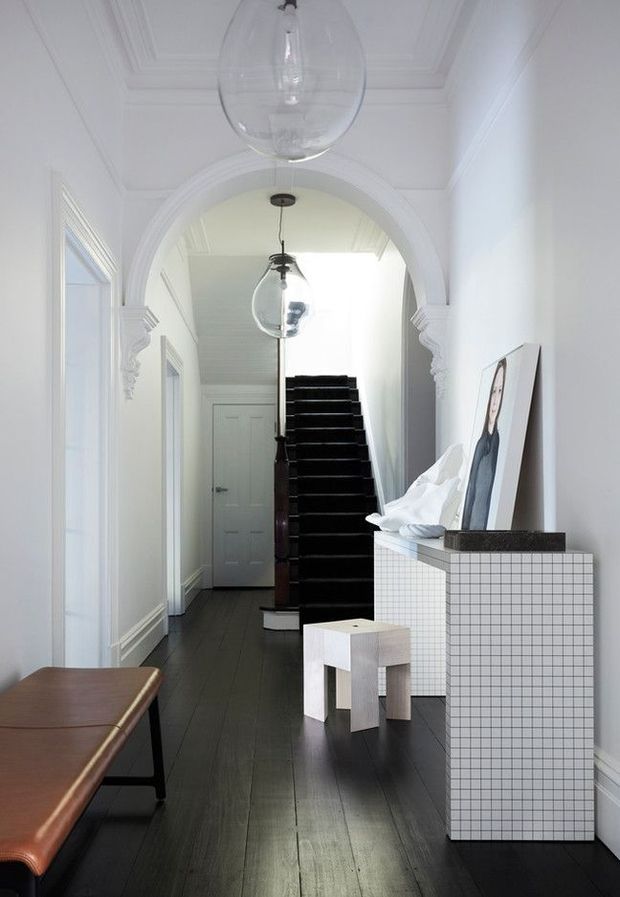 A bright, narrow residential hallway or entryway with white walls, ornate arched opening and decorative ceiling mouldings. Dark timber flooring leads to a black staircase, while a white gridded-tile console stands on the right with framed artwork and a small pale stool. Glass pendant lights hang along the circulation space, and a brown upholstered bench occupies the foreground.