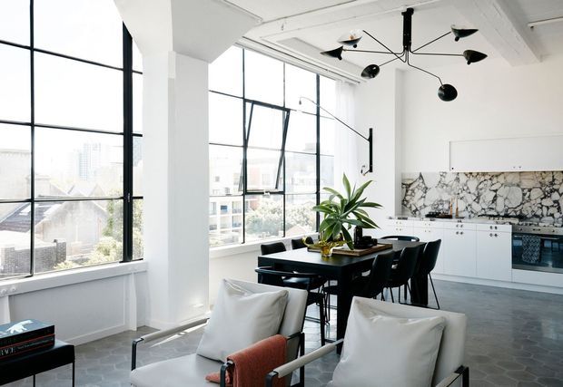 Bright open-plan loft interior with large black-framed industrial windows, white structural columns and ceiling, a black dining table with chairs, a compact white kitchen, marble-look splashback, and a seating area in the foreground. A sculptural black pendant light and indoor plants add contemporary character, with an urban city view beyond the glazing.