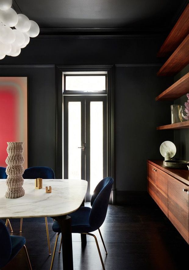 A dramatic, refined dining room with black-painted walls, ceiling, trim and dark flooring. A white marble-topped dining table is surrounded by deep blue upholstered chairs, while a long built-in dark timber sideboard with open shelves occupies the right wall. A tall glazed door provides strong backlighting, and a sculptural clustered white pendant hangs overhead. Warm timber shelving and cabinetry contrast with the dark interior.