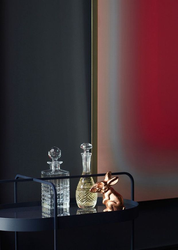 A moody, highly styled interior vignette featuring a black circular bar cart or drinks trolley with two cut-crystal decanters and a small metallic rabbit sculpture. The background combines a near-black wall with a narrow reflective vertical panel and a deep red illuminated wall, creating a dramatic, luxurious atmosphere.