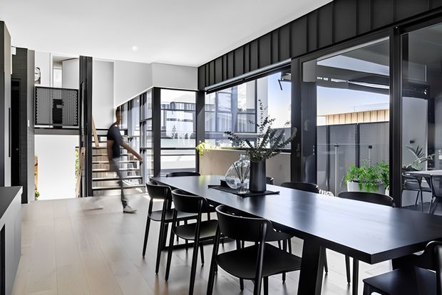 A bright, open-plan contemporary dining area with a long black dining table and matching black chairs. Full-height black-framed glazing and glazed internal partitions create strong visual connections to the exterior and adjacent spaces. A modern staircase with open risers and black balustrade rises at the rear, while dark vertical wall cladding contrasts with pale flooring and white walls.