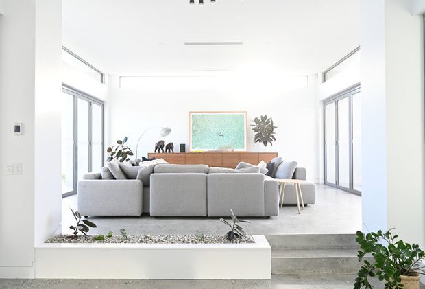 Bright, airy contemporary living room with a large grey sectional sofa facing a low timber media console and framed artwork. White walls and ceiling, extensive floor-to-ceiling glazing, polished concrete flooring, indoor plants, and a split-level step create a clean, minimalist interior with strong natural light.