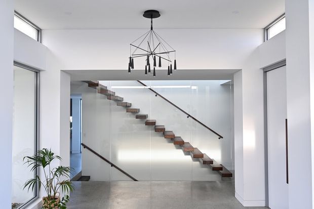 Bright contemporary interior centered on a floating staircase with timber treads, slim dark metal handrails, and a glass balustrade. White walls, polished concrete flooring, high-level glazing, and a sculptural black pendant create a clean, open stair hall.