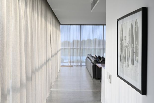 A bright, minimalist interior corridor or entry passage leading into a living room. Full-height sheer curtains line the left side, while a large black-framed artwork and light switches occupy the right wall. A dark sofa is visible ahead beside floor-to-ceiling glazing, with a pale smooth floor and clean white walls creating an airy, contemporary feel.