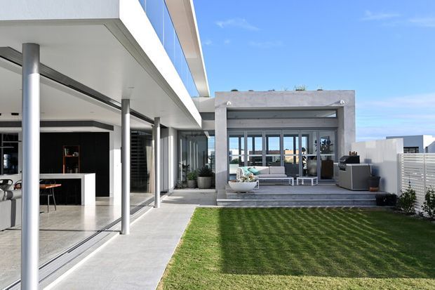 Contemporary residential courtyard viewed from the garden, featuring a sleek white and grey flat-roofed structure with extensive floor-to-ceiling glazing, slender metal columns, polished concrete paving, and a covered outdoor lounge. The open-plan interior kitchen and dining area is visible through the glazing, while a manicured lawn and privacy fencing occupy the foreground.