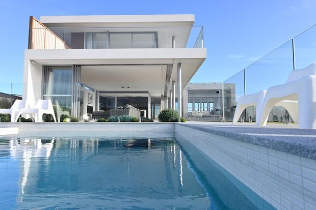 Bright contemporary multi-level white residence viewed from the swimming pool, with expansive floor-to-ceiling glazing, projecting flat rooflines, glass balcony balustrades, and a covered outdoor living area. The pool and pale tiled coping dominate the foreground, while sculptural white outdoor furniture and glass pool fencing frame the scene.