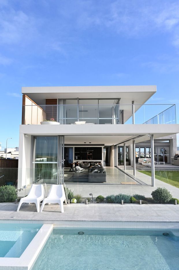 A bright modern two-storey residence viewed from the pool courtyard. The home has crisp white rendered walls, broad flat roof planes, extensive floor-to-ceiling glazing, glass balustrades, and a covered open-plan ground-floor living area opening onto a paved terrace. A swimming pool occupies the foreground, with minimalist landscaping and outdoor seating.