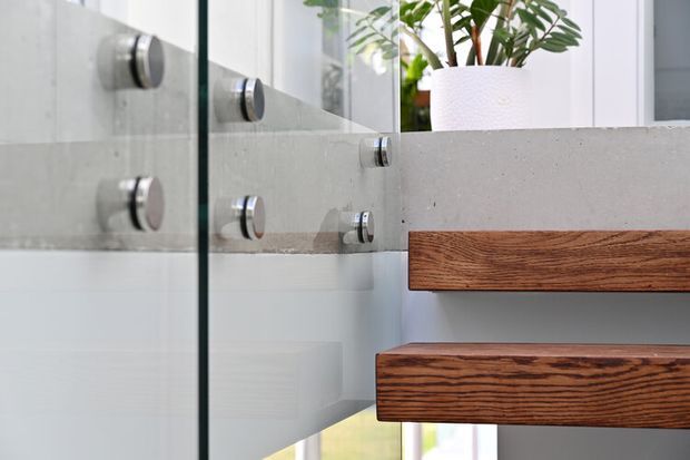 Close-up architectural detail of a contemporary stairway beside a glass balustrade. Thick dark timber stair treads project from a smooth light-grey concrete or rendered wall, with circular metal fixings visible through the glass. A potted plant and bright glazing appear in the background.
