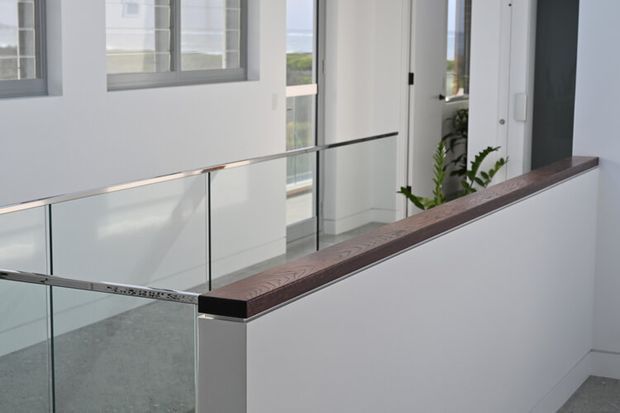 Bright contemporary interior landing or stairwell with white walls, large windows, a frameless glass balustrade with slim metal handrail, and a dark timber handrail cap along a half-height partition. A glimpse of greenery and exterior landscape is visible through the glazing.
