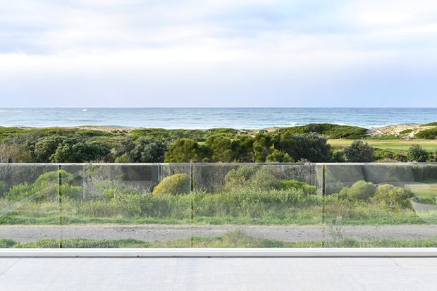 A wide coastal view seen from behind floor-to-ceiling glazing, with a calm ocean horizon, sandy dunes, native coastal vegetation, and a transparent glass balustrade or barrier in the foreground. The image is bright and minimal, with a pale paved or tiled surface at the bottom edge and no visible interior furnishings.