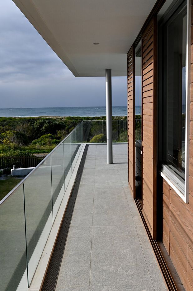 A contemporary coastal balcony or terrace viewed alongside a house facade. The space features large grey tiled paving, frameless glass balustrades, a slim cylindrical metal column, a broad white soffit with recessed downlights, and warm horizontal timber wall cladding with dark-framed glazing. Dense coastal vegetation and the ocean are visible beyond the terrace.