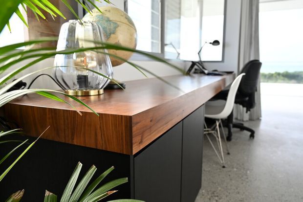 A contemporary home office with a large dark built-in desk featuring a warm timber benchtop and matching timber front, black cabinetry below, and two ergonomic office chairs. Floor-to-ceiling glazing, sheer curtains, a globe and desk lamp are visible, with indoor tropical plants adding a natural element.