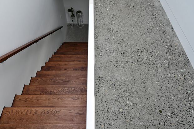 Interior view looking down a contemporary stairway with warm dark timber treads, a simple white wall, and a slim timber handrail. A contrasting grey terrazzo-like surface forms the stair-side edge or adjacent landing, with a small plant arrangement visible below.