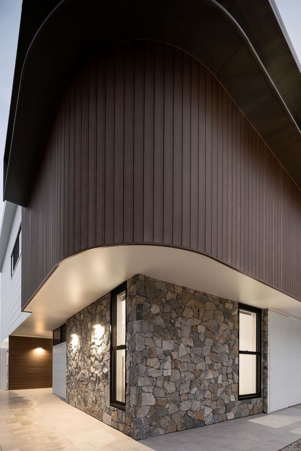 Modern residential exterior viewed from a covered entry or driveway, featuring a dramatic curved dark roof canopy, vertically ribbed dark timber cladding, a natural irregular stone feature wall, large black-framed windows, and pale paved surfaces. Warm architectural lighting highlights the stone facade and sheltered approach.