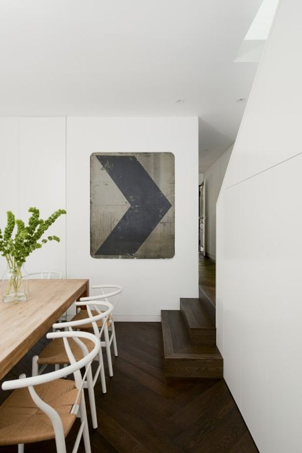 A bright, minimalist dining area with a light timber dining table and white-framed woven chairs beside a compact dark timber stairway. White built-in walls and cabinetry create a clean backdrop, with a large monochrome arrow artwork and a vase of greenery adding visual interest. A roof skylight or clerestory opening brings natural light into the space.