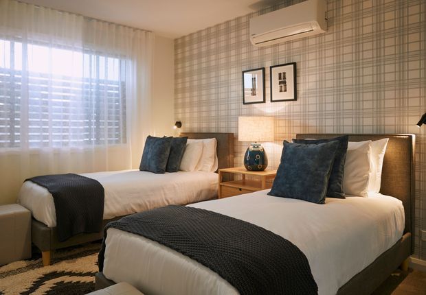 A bright, neatly styled bedroom with two beds, white bedding, dark charcoal textured throws, and deep blue-grey cushions. A warm timber bedside table sits between the beds, with a woven-pattern feature wall, framed artwork, bedside lighting, and a wall-mounted air conditioner. Sheer curtains and horizontal blinds filter daylight through the large window.