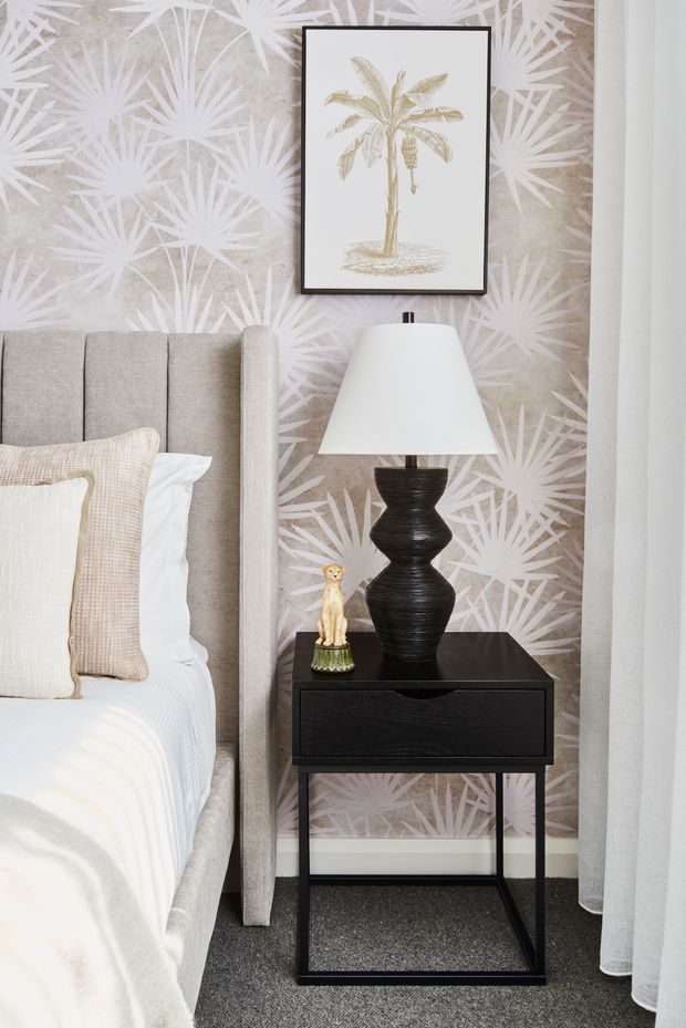 A refined bedroom vignette showing a tall upholstered bedhead beside a slim black bedside table with a sculptural black lamp, framed botanical artwork, and a small decorative figurine. The wall is covered in a pale botanical palm-patterned wallpaper, with layered neutral bedding and sheer white curtains. The overall composition is elegant, calm, and softly contemporary with tropical-inspired detailing.