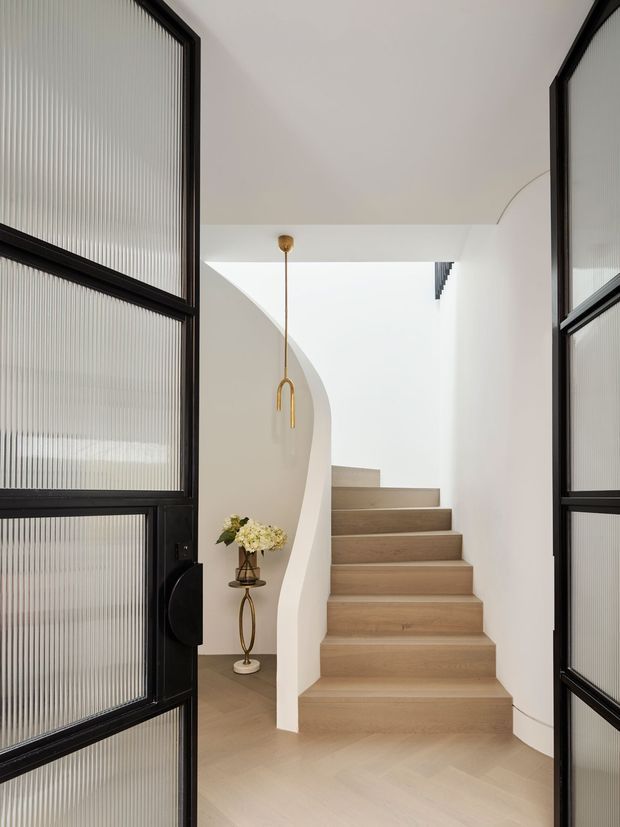 A refined residential stairway viewed through open black-framed fluted-glass doors. The space features a sculptural curved white stair wall, pale timber stair treads and herringbone flooring, a slim brass pendant light, and a small decorative floral arrangement beneath the stairs. The palette is light and neutral with strong black-framed glazing accents.