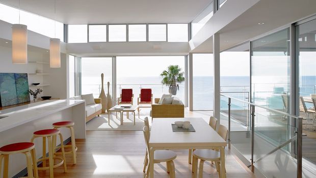Bright open-plan coastal residence with a combined kitchen, dining and living area opening onto a glazed balcony or terrace. The room features white cabinetry and surfaces, pale timber flooring, a white dining table with sculptural timber stools, a light sofa, large sliding glass doors, clerestory glazing and ocean views. The interior is airy and contemporary with strong indoor-outdoor connection.