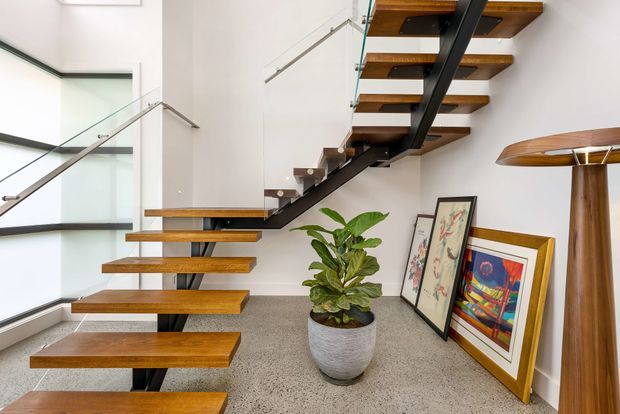 Bright contemporary stairway with floating timber treads supported by a dark central steel spine, clear glass balustrades, and white walls. A terrazzo-like floor, potted plant, framed artwork, and a sculptural timber floor lamp complete the open circulation space.