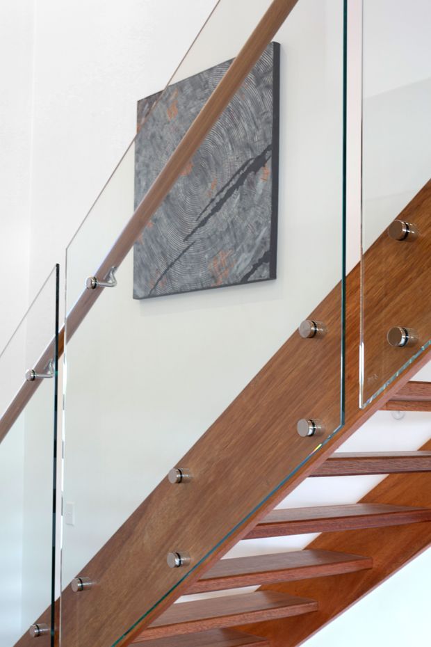 Close-up interior view of a contemporary open-riser staircase. The stair has warm timber treads and a broad timber stringer/side structure, paired with clear glass balustrade panels secured by metal standoffs and a timber handrail. White walls and a dark abstract artwork form the background.