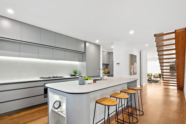 Bright contemporary kitchen with a large island featuring an integrated sink and waterfall-style light benchtop, grey handleless cabinetry, a pale stone splashback, timber flooring, and timber-and-metal bar stools. A timber open-riser stair or screen structure is visible to the right, with a living area beyond.