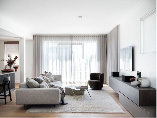 Bright contemporary living room with a large grey sectional sofa, sculptural armchair, round coffee table, pale area rug, and low dark wood-and-grey media console. Full-height sheer curtains cover glazed doors or windows at the rear, while a dining area is visible to the left. The room has white walls and ceiling, light timber flooring, and restrained neutral styling.