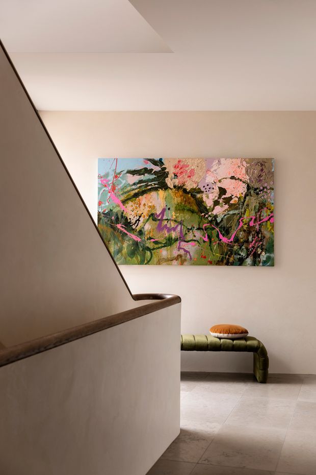 A softly lit residential stair landing or hallway with warm beige plaster walls, a sculptural stair balustrade, pale stone or tile flooring, and a low olive-green curved bench with a rust-orange cushion. A large vibrant abstract floral painting provides the main decorative focal point.