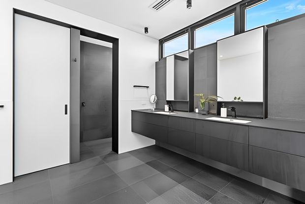 A contemporary bathroom with a long floating dark vanity, two integrated basins, wall-mounted black tapware, large frameless mirrors, and a separate enclosed shower area. The room has large dark grey floor tiles, white walls, charcoal grey cabinetry and panels, black-framed clerestory windows, and minimalist fittings. Bright natural light enters through the upper glazing, creating a clean, sophisticated monochrome interior.