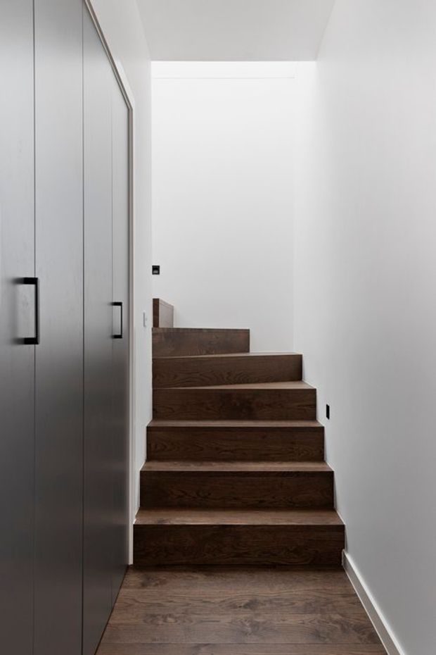 Minimalist interior stairway with dark timber treads and risers, white walls and ceiling, and a tall flush built-in grey cabinet with black handles along the left side. The compact staircase rises toward a bright landing, creating a clean, understated modern composition.