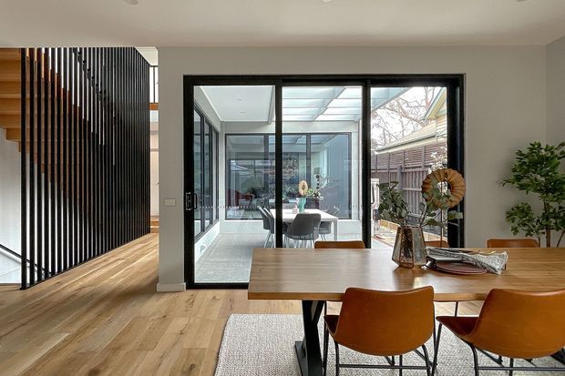 A contemporary dining area with a timber dining table and warm tan upholstered chairs, opening through black-framed glazing to a bright enclosed courtyard or sunroom. Light timber flooring, neutral walls, a vertical timber-slat screen, and indoor plants create a warm, refined interior. A partial stair and circulation space are visible to the left.