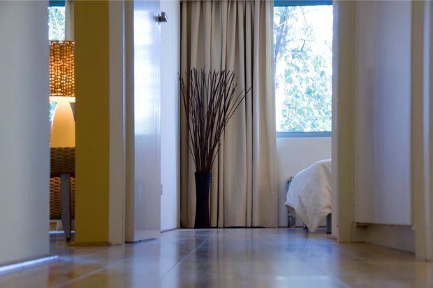 View down a polished light-toned hallway toward a bedroom, with cream curtains, a tall decorative branch arrangement, white doors and trim, and glimpses of a warm-lit adjoining space and bed. The interior is bright, clean, and contemporary with strong linear perspective.