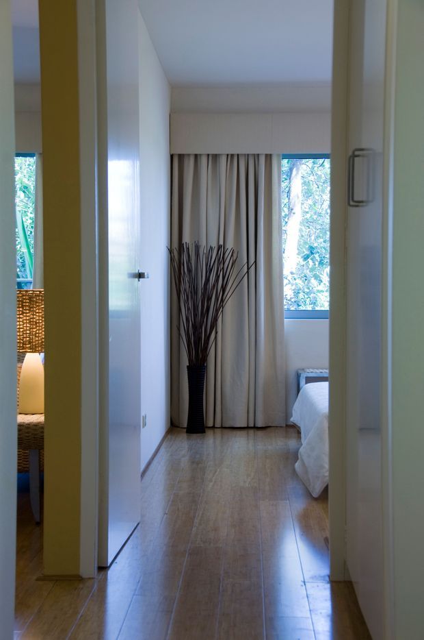 View through a hallway toward a bedroom, with warm timber flooring, white walls and doors, full-height cream curtains, and a window overlooking greenery. Part of a bed is visible in the bedroom, while a woven table lamp appears in an adjacent room on the left.