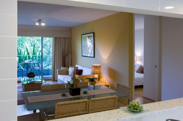 View from a kitchen through a wide opening into an open-plan dining and living area. The dining zone has a glass-topped rectangular table with woven wicker chairs, while the living area includes a white sofa, patterned rug, warm yellow feature wall, framed artwork, and a large glazed opening to a leafy balcony or exterior. A bedroom is partially visible through a doorway at right. White kitchen cabinetry and a pale speckled benchtop frame the foreground.